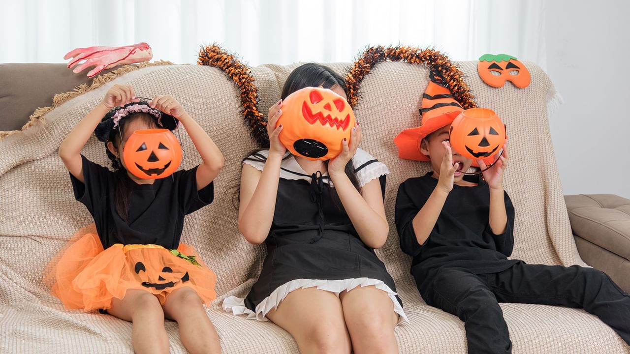 Three kids in Halloween costumes holding pumpkin masks sit on a couch.