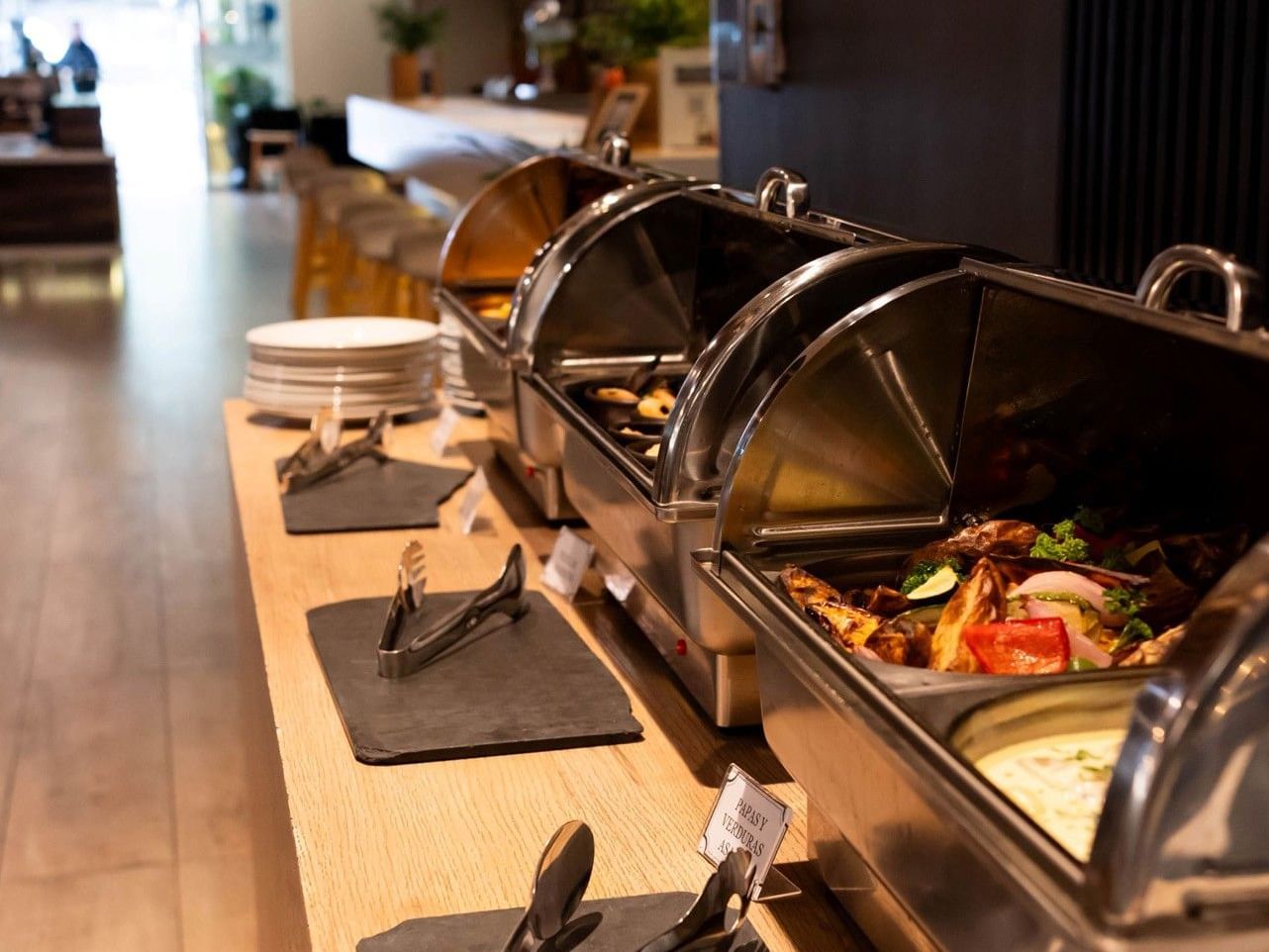 Mesa de bufé con recipientes para la comida y cubiertos en el hotel HCC Costanera Concepción.