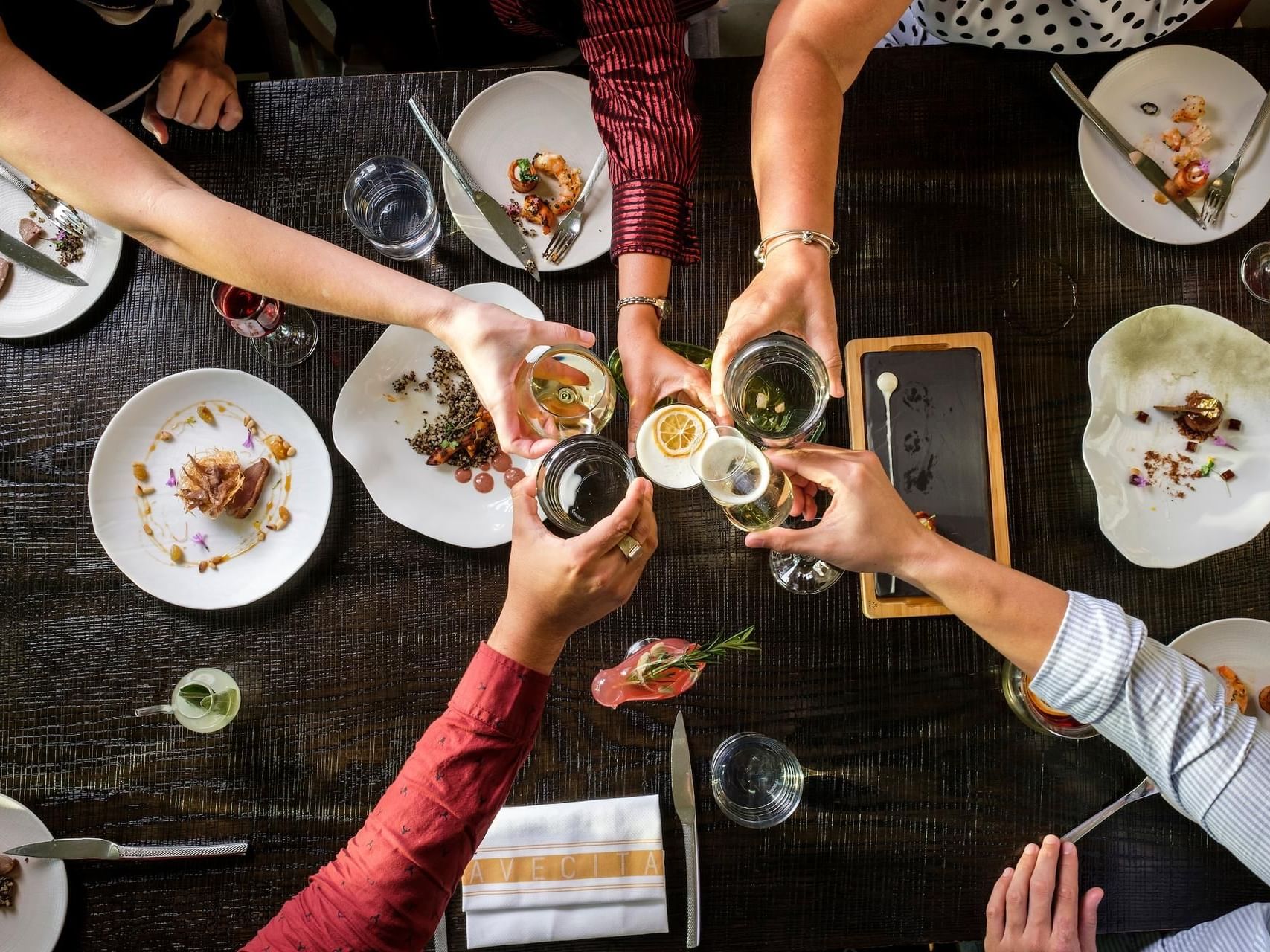 Group of hands toasting with drinks over an set dining table with dishes in Avecita at The Residences at Seafire.