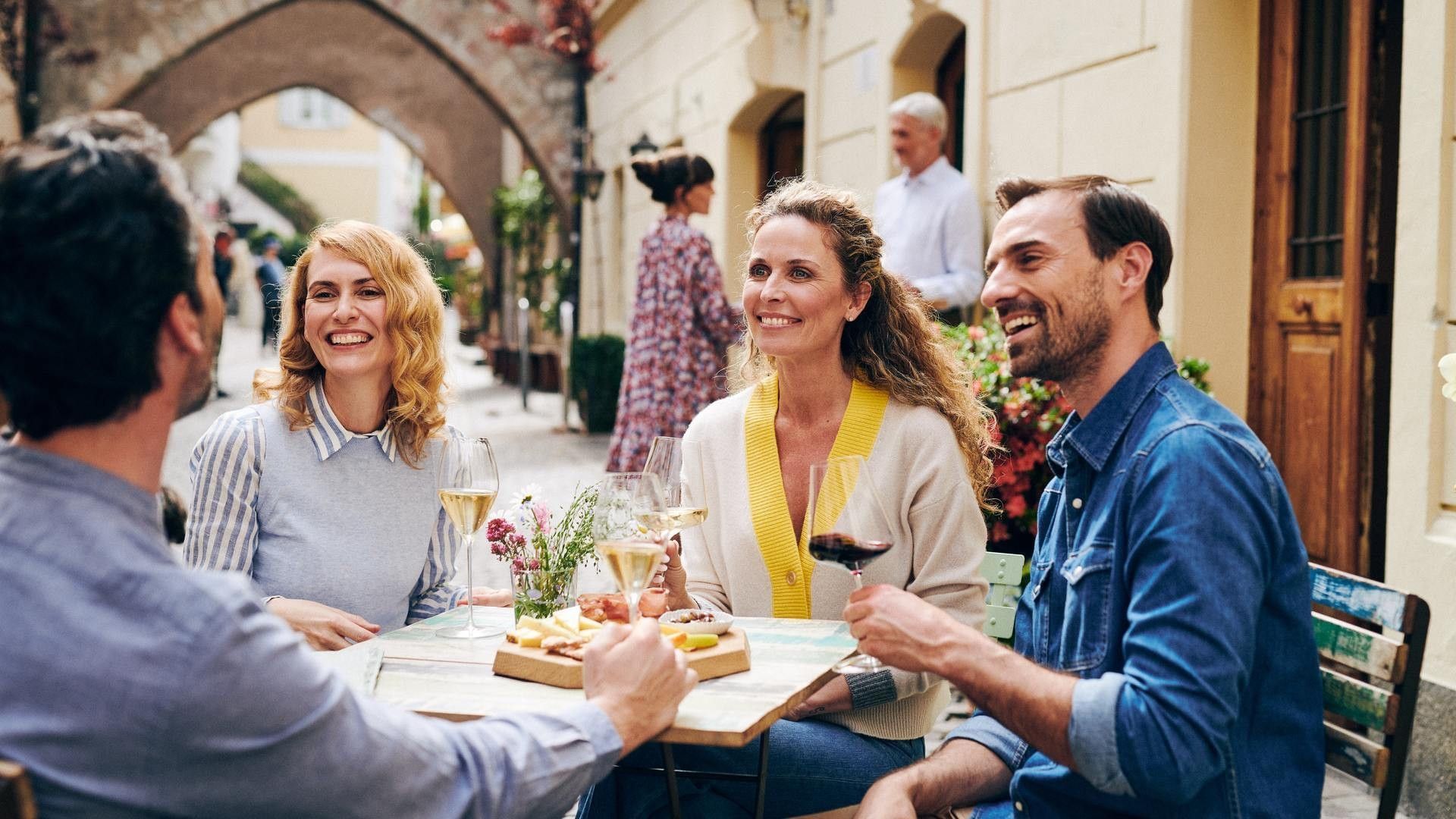 Quattro persone sorridono e ridono mentre bevono vino e mangiano cibo seduti a un tavolo all'aperto.