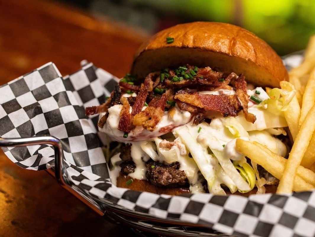 Gourmet burger topped with bacon, coleslaw, and sauce served with fries in checkered basket at The Burger Dive