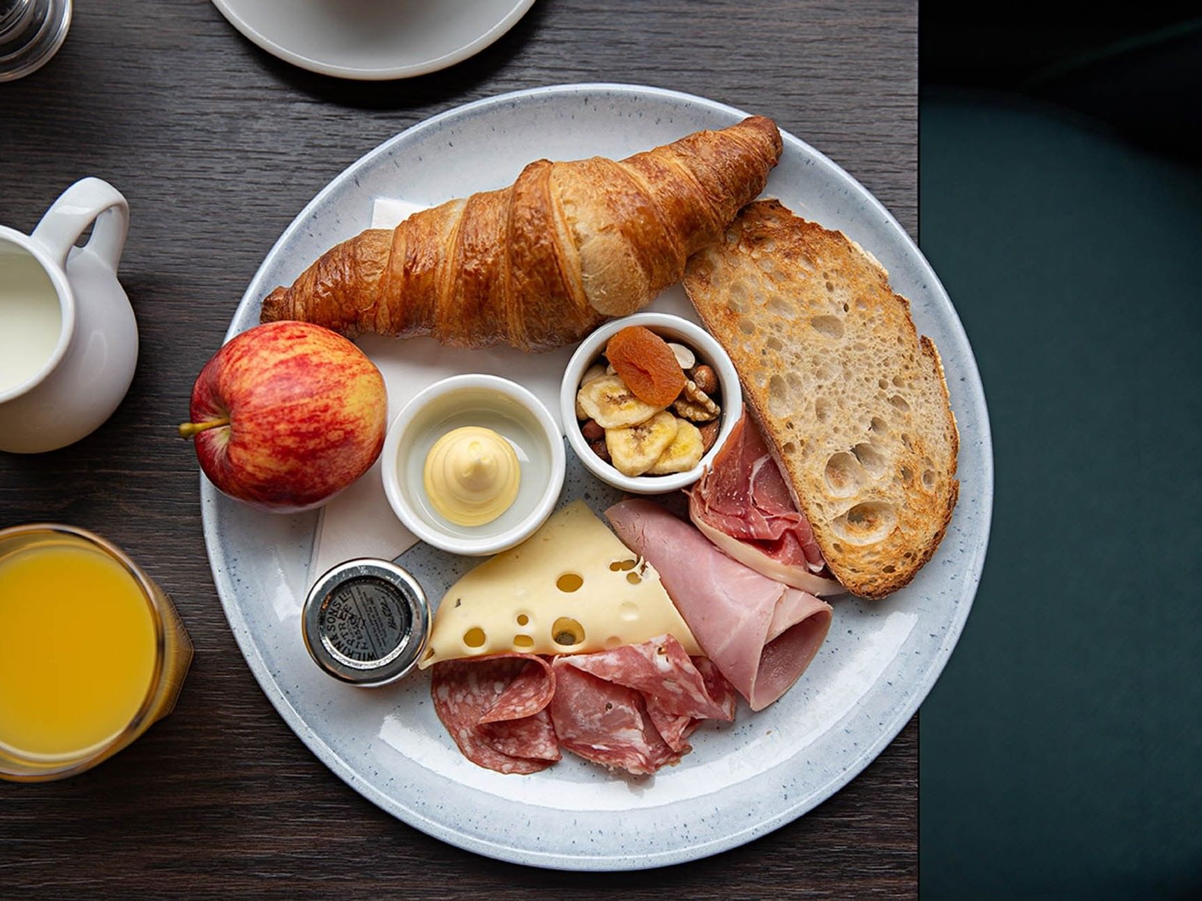 Plate with croissant, apple, bread, cheese, meat, fruit, and nuts, with milk, juice, and condiments nearby.