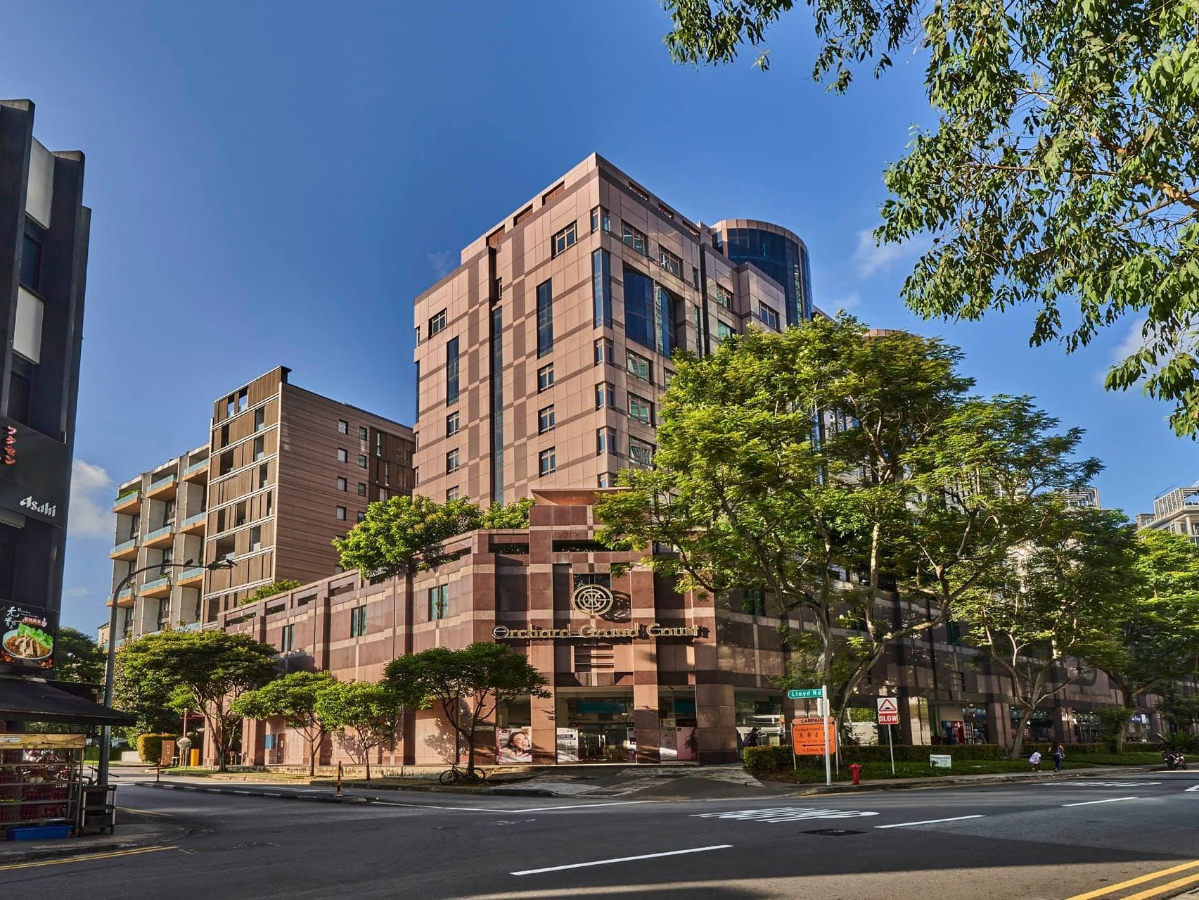 Modern exterior view of Orchard Grand Court, framed by trees, shops and street view under a clear blue sky