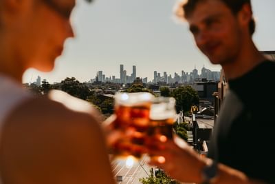 Couple clinking glasses in the foreground with the city skyline in the background at Amora Herencia Riverwalk Melbourne