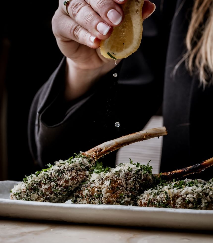 Hand squeezes a lemon slice over three herb-crusted lamb chops on a plate at The Kitchen