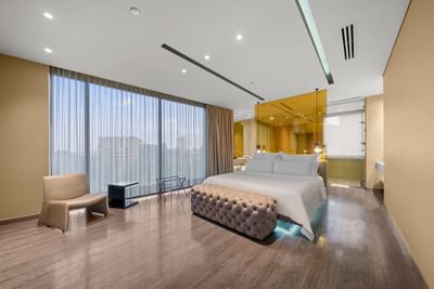 Habitación moderna con cama grande, silla, banco, cortinas grandes y baño de vidrio en esquina.