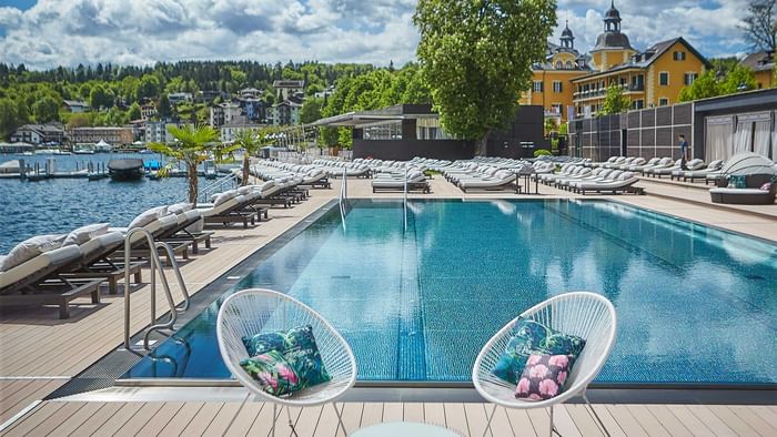 Blick auf einen großen Pool, umgeben von Liegen und Gebäuden mit einem See im Hintergrund.