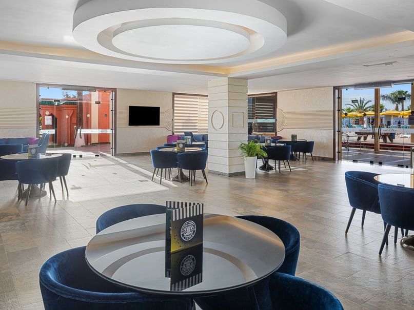 Modern lounge area with blue chairs, round tables, and large glass doors showing outdoor seating and umbrellas.