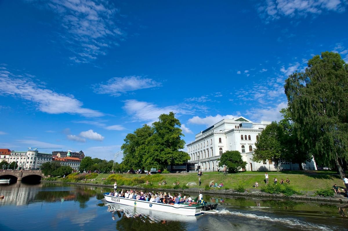A boat filled with people sails on a river near a park with trees and a large white building.