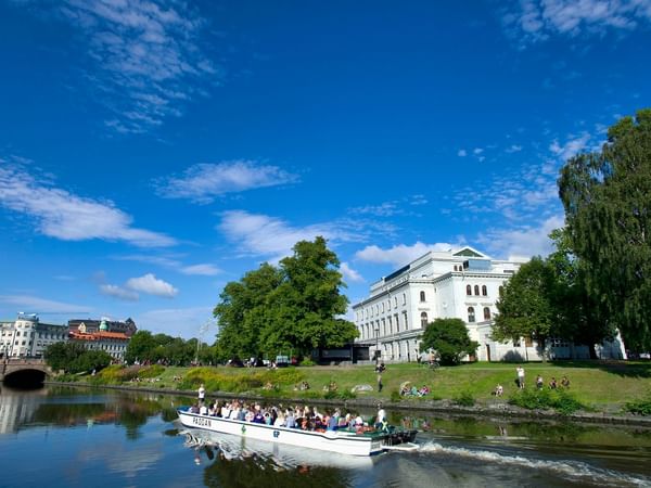 White boat moving on a canal with buildings and trees in Gothenburg, close to Hotel Riverton, Ideal for summer coolcation.