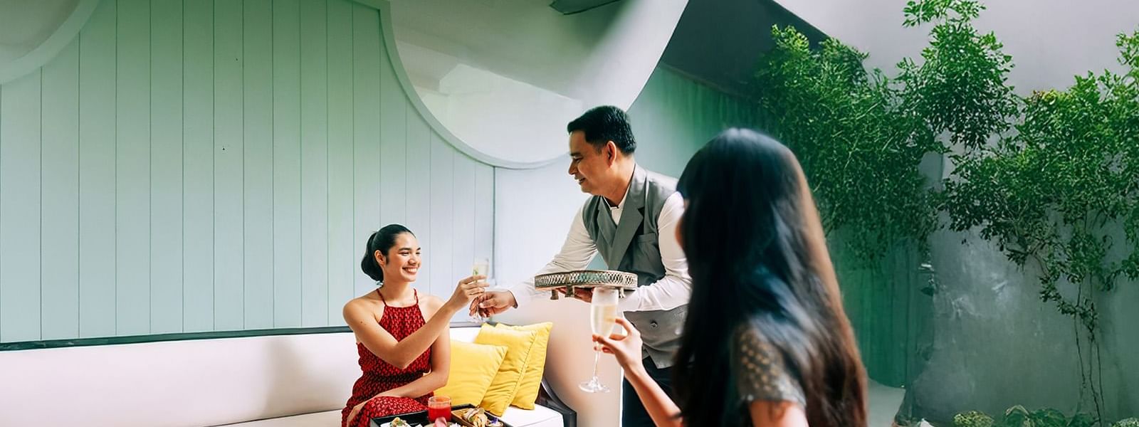 Two women in red dresses toast with champagne as a waiter presents a tray of desserts.