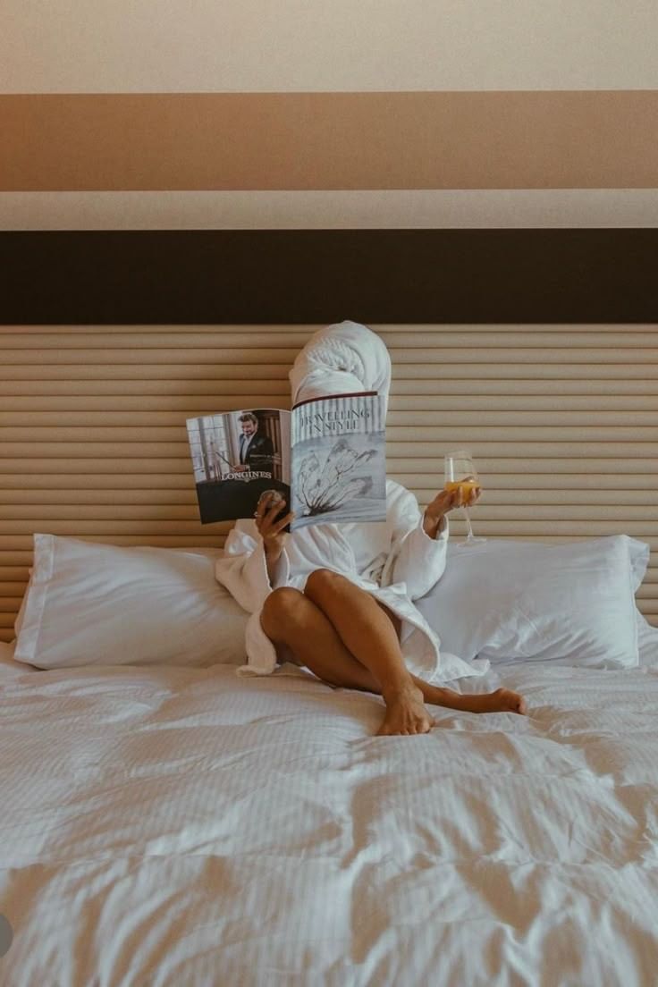 A person in a white robe sits on a bed, reading a magazine and holding a champagne flute.