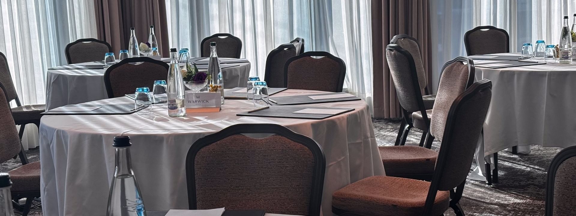 Round tables with white linens by brown chairs under sheer curtains in Rigi + Cervin at Warwick Geneva