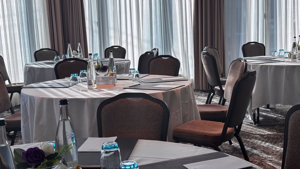 Round tables with white linens by brown chairs under sheer curtains in Rigi + Cervin at Warwick Geneva