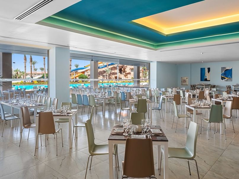 Large dining area with many tables and chairs, blue and yellow ceiling, and outdoor pool view.