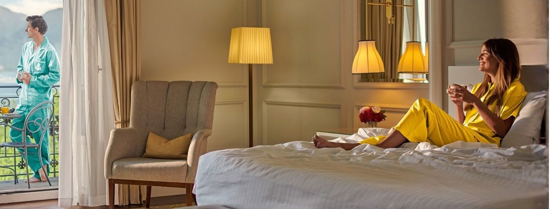Woman in yellow loungewear relaxing on a bed in a hotel room with a balcony and scenic view.