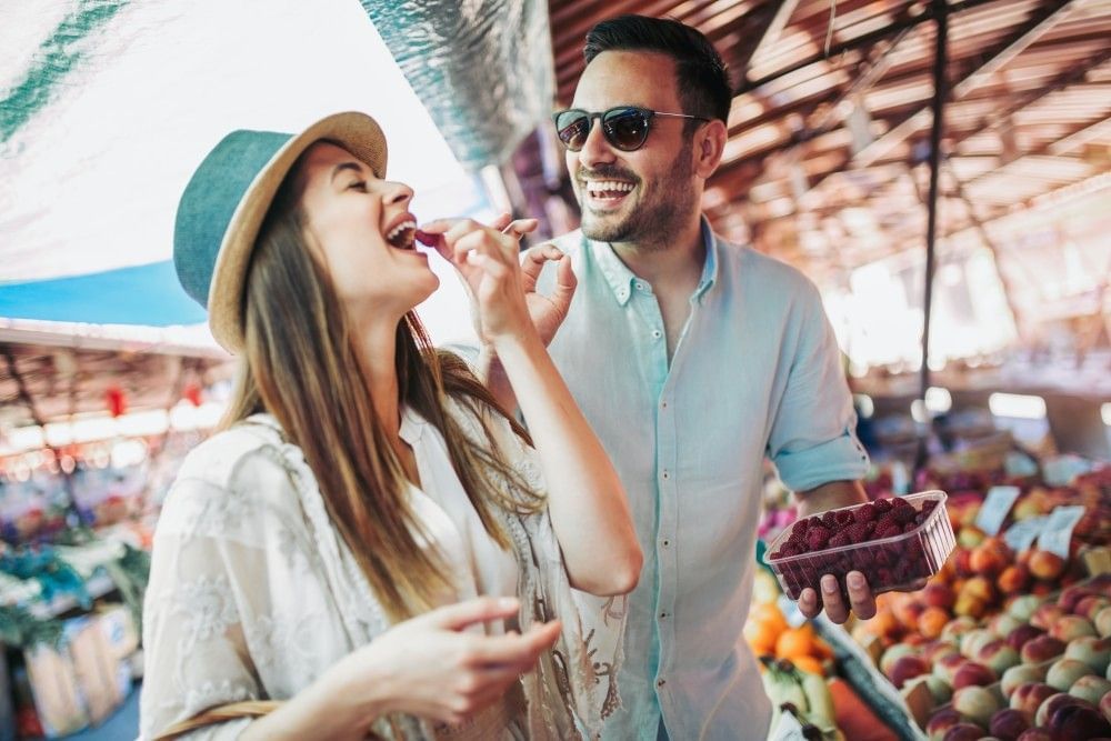 A man and woman playfully eat berries while laughing together at a farmer's market stand.