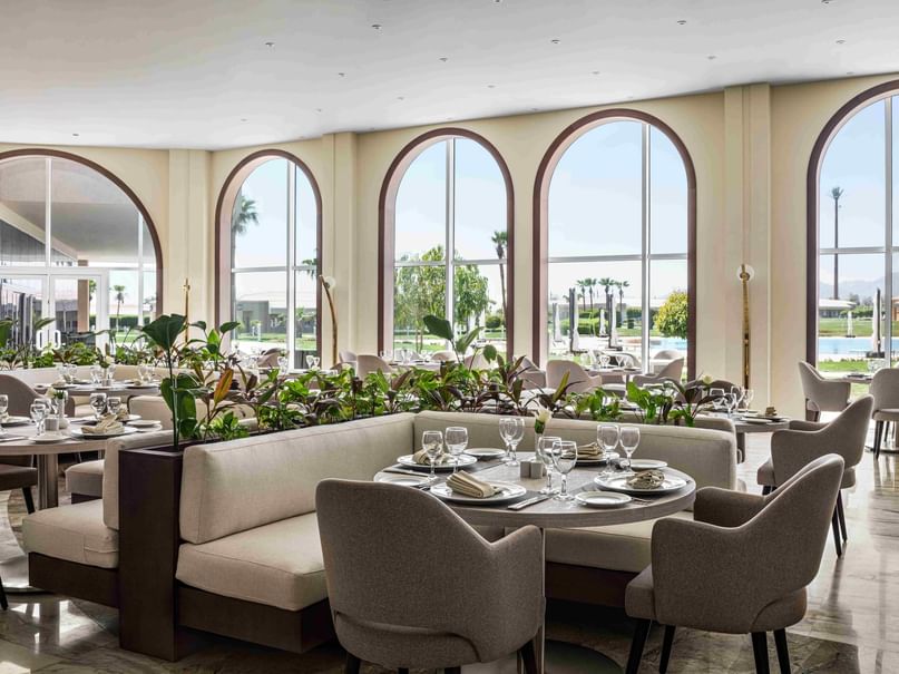 An elegant restaurant with arched windows, tables set with fine dining items, and a view of greenery.