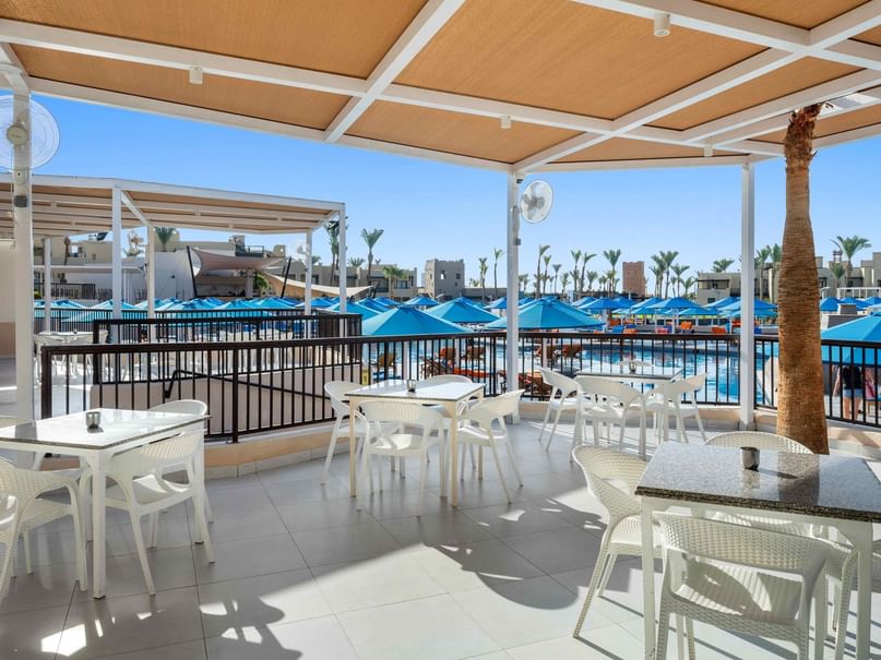 Outdoor patio with white tables and chairs, pool with blue umbrellas in background, and a clear blue sky.