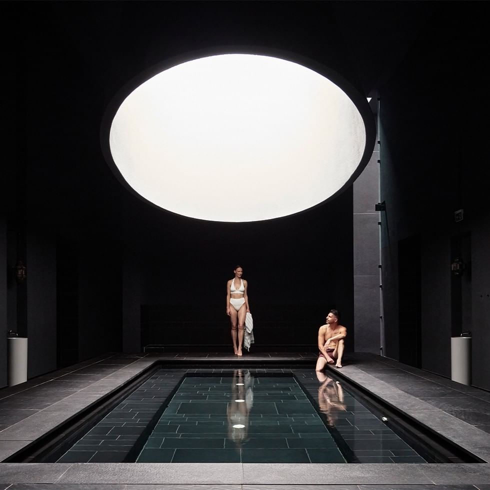 Two people by a pool under a large circular skylight in a dark room.