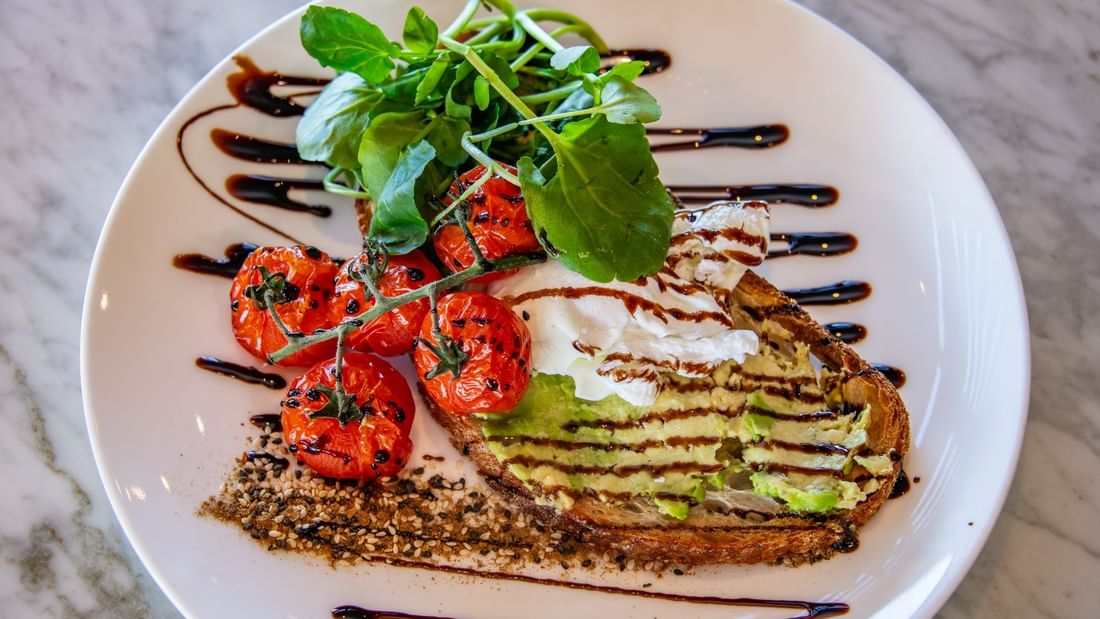 A plate with avocado toast, cherry tomatoes, and greens, drizzled with dark sauce on a marble surface.