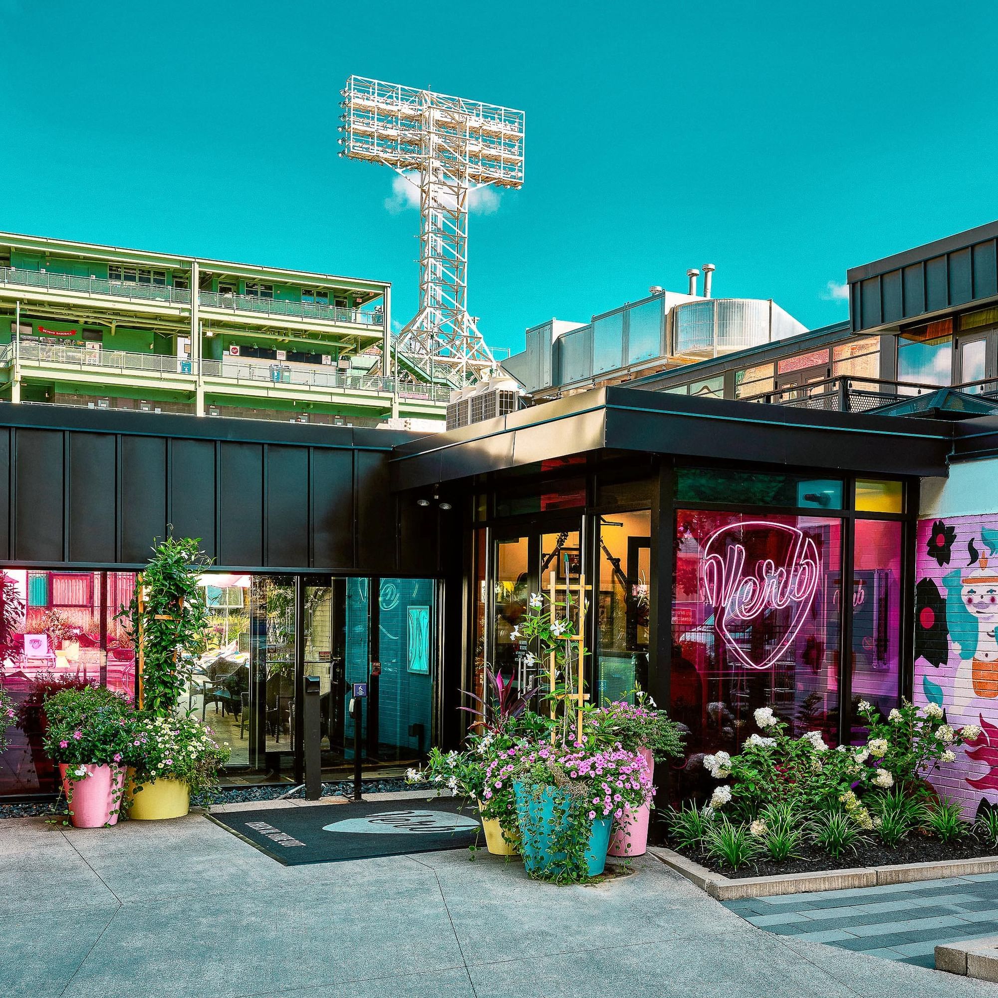 Modern hotel exterior with colorful neon signage, vibrant planters, and glass facades under a bright blue sky in Boston.
