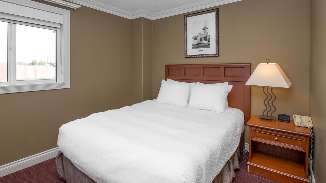 Guest room with white bedding, wooden headboard, nightstand with lamp, and framed artwork at Coast Edson Hotel.