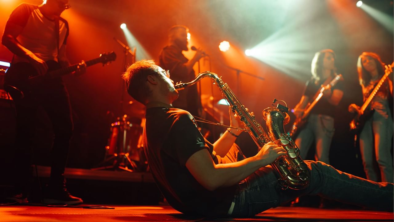 A man plays saxophone on stage with a band, under red and green spotlights.