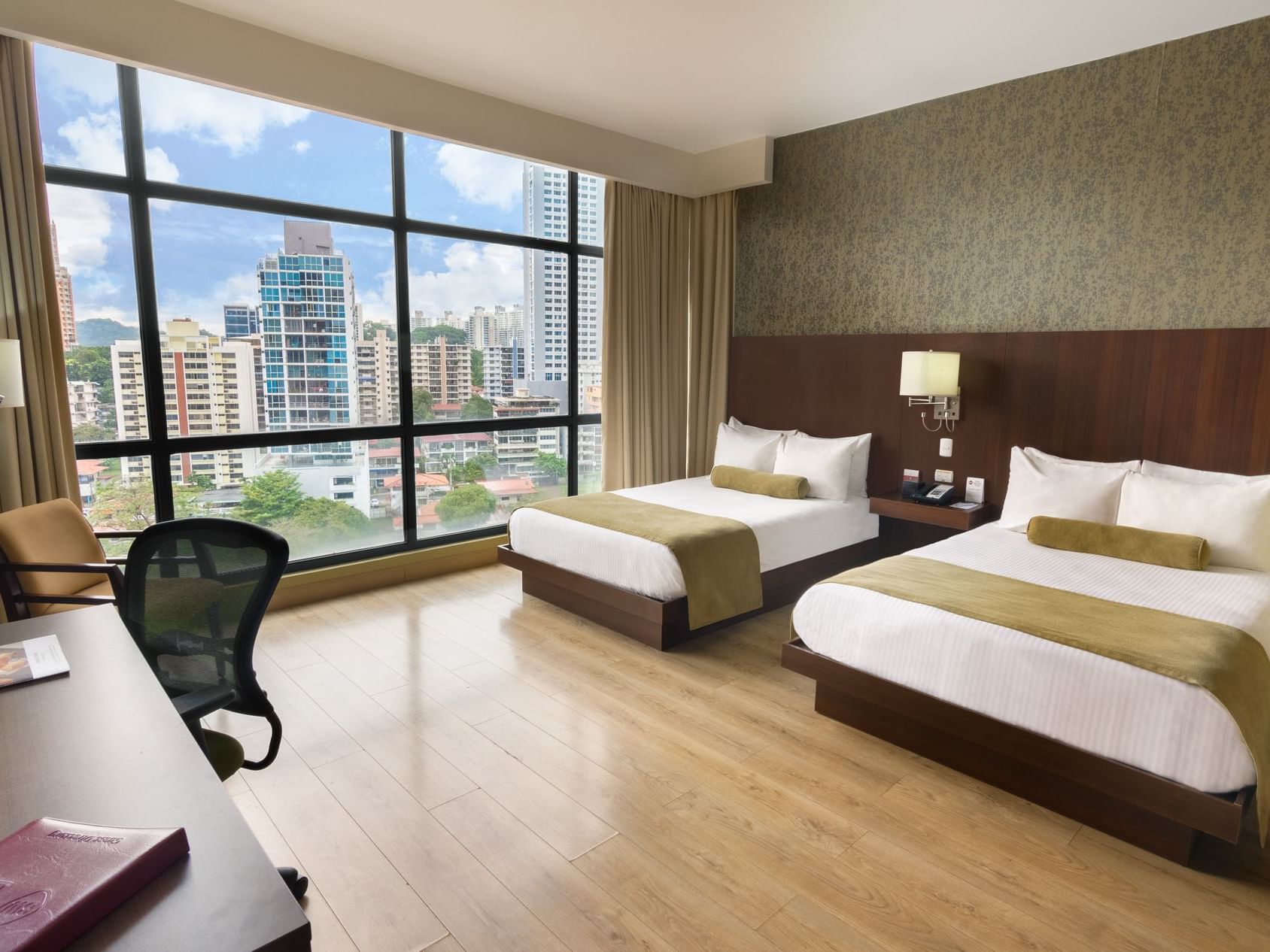 The Superior Twin Room with two double beds, a desk, a chair, a side table in between the beds with a lamp and a view of the city