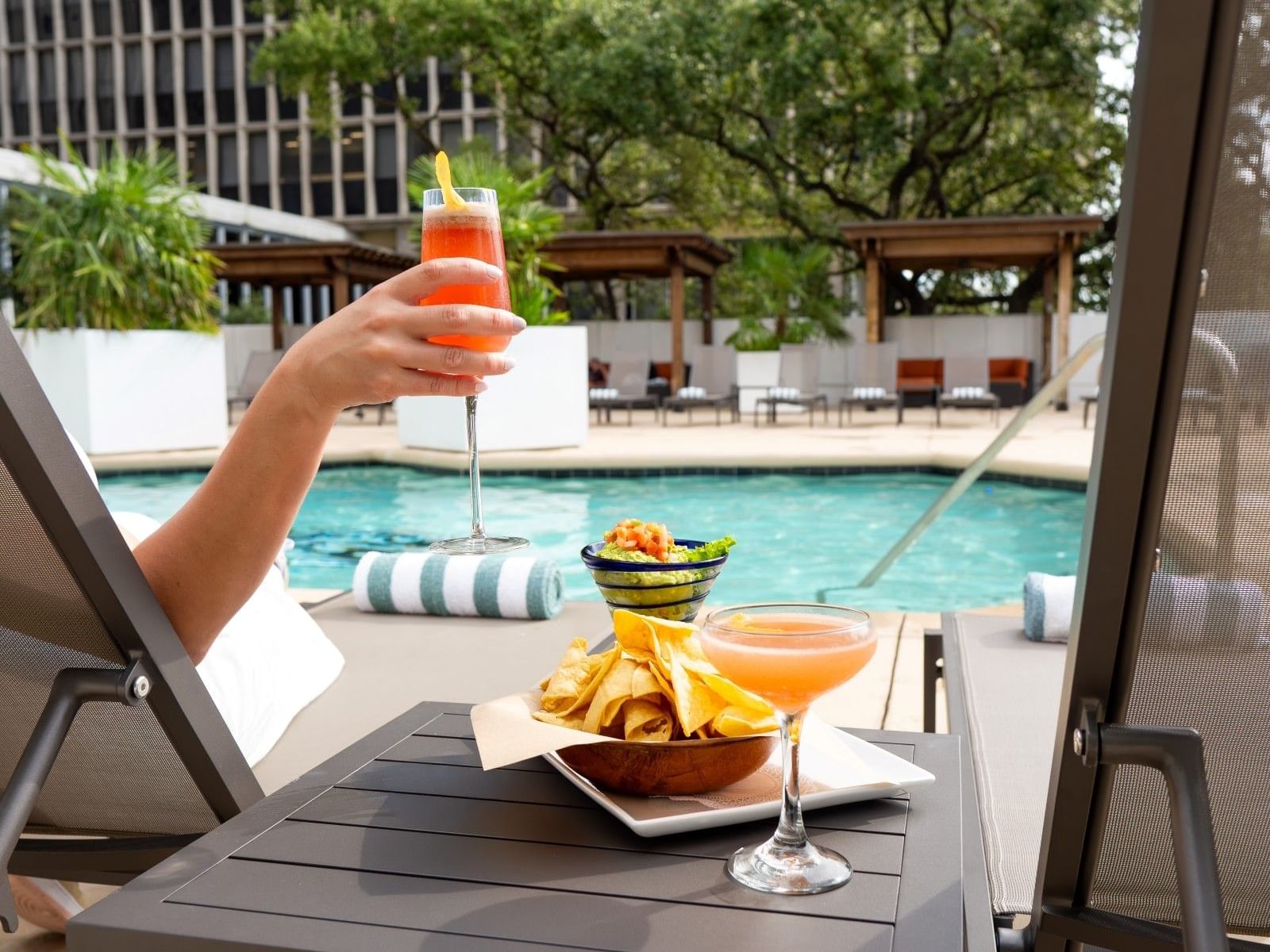 Hand holding a cocktail next to a table with a plate of chips and another cocktail.