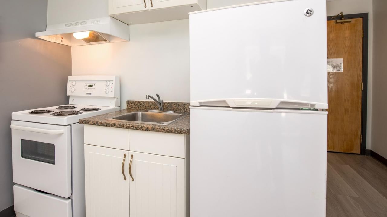 White kitchen with stove, refrigerator, and cabinets, with wooden door on the right.