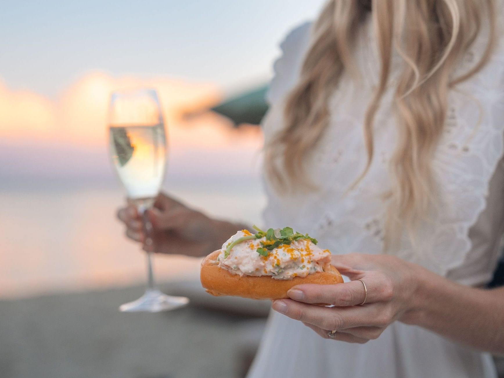 Woman in white dress holding gourmet appetizer on bread with glass of white wine at sunset by the water
