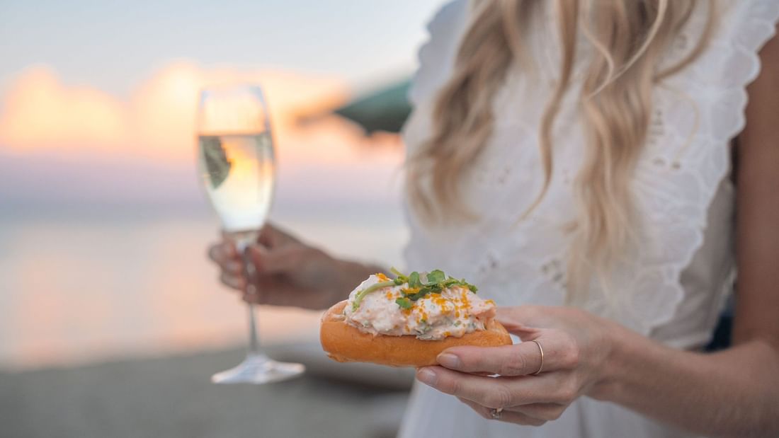 Woman in white dress holding gourmet appetizer on bread with glass of white wine at sunset by the water