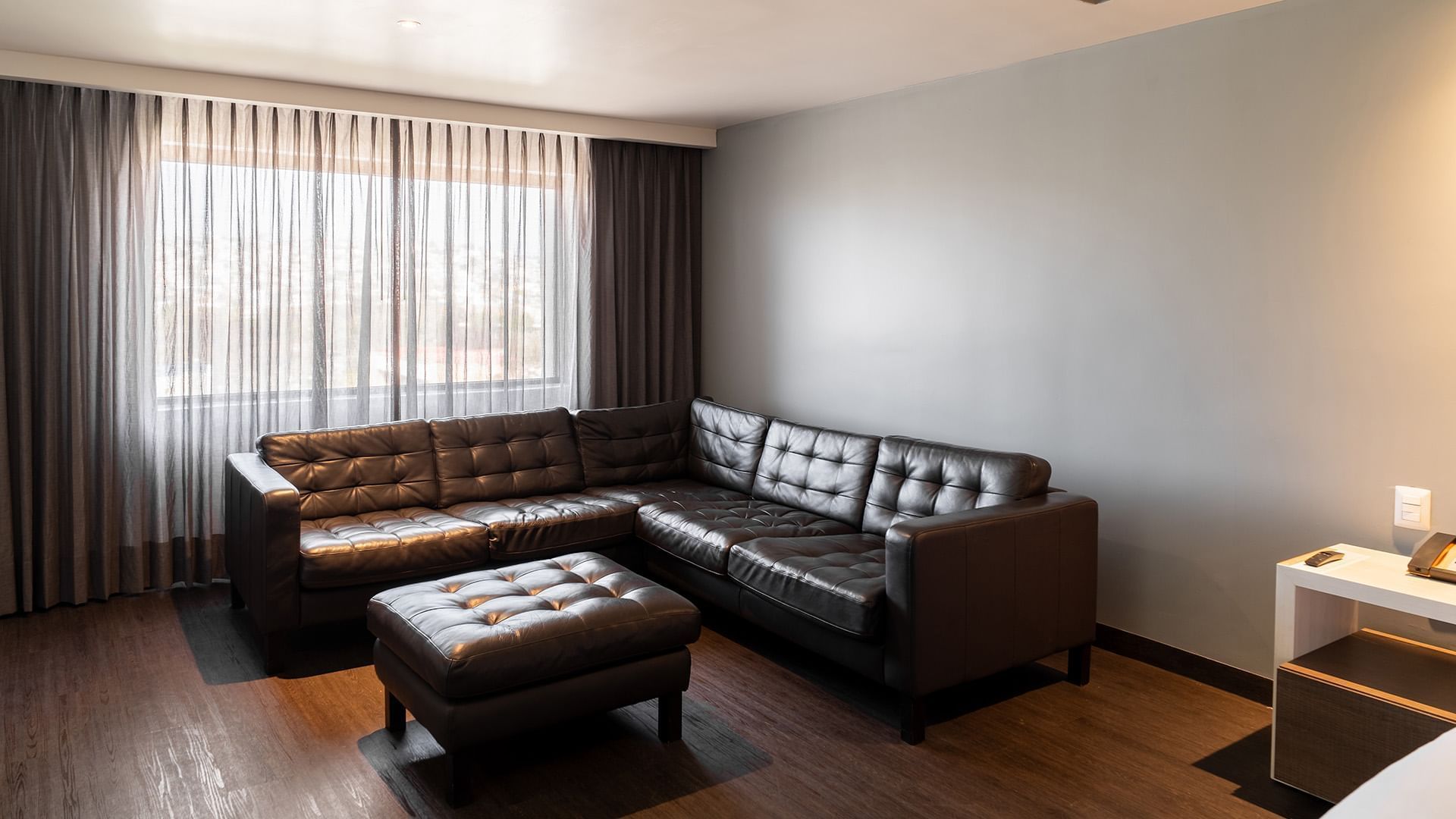 King Junior Suite featuring a dark L-shaped leather sofa, matching ottoman, and curtains at Real Inn Tijuana