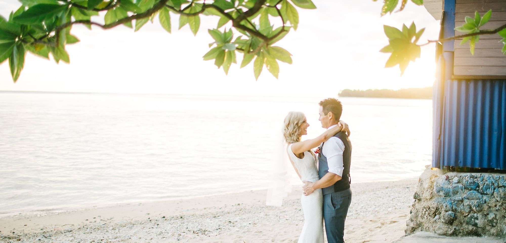 Wedding couple by the shore under green branches near a blue wood building at Warwick Fiji Resort and Spa
