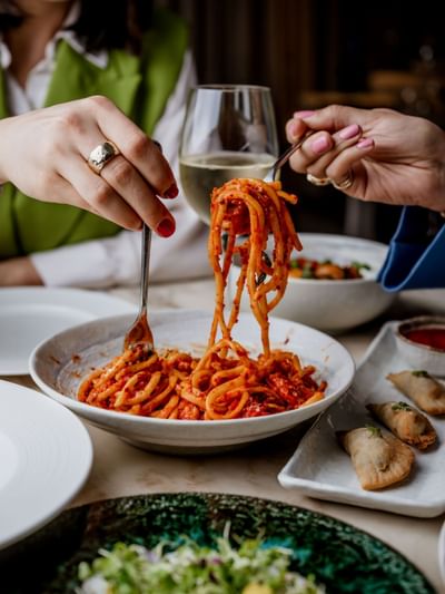 Two hands twirl pasta from a bowl, accompanied by a glass of white wine on a table at The Kitchens