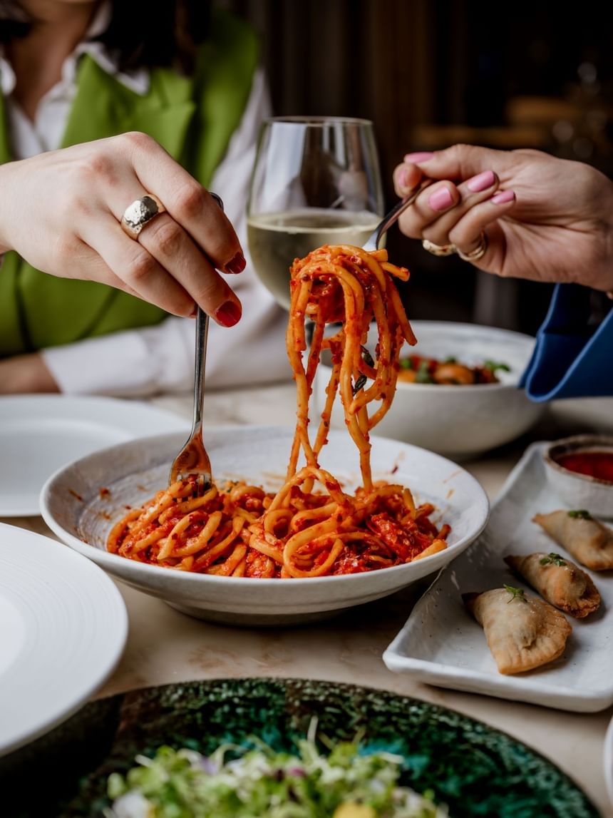 Two hands twirl pasta from a bowl, accompanied by a glass of white wine on a table at The Kitchens