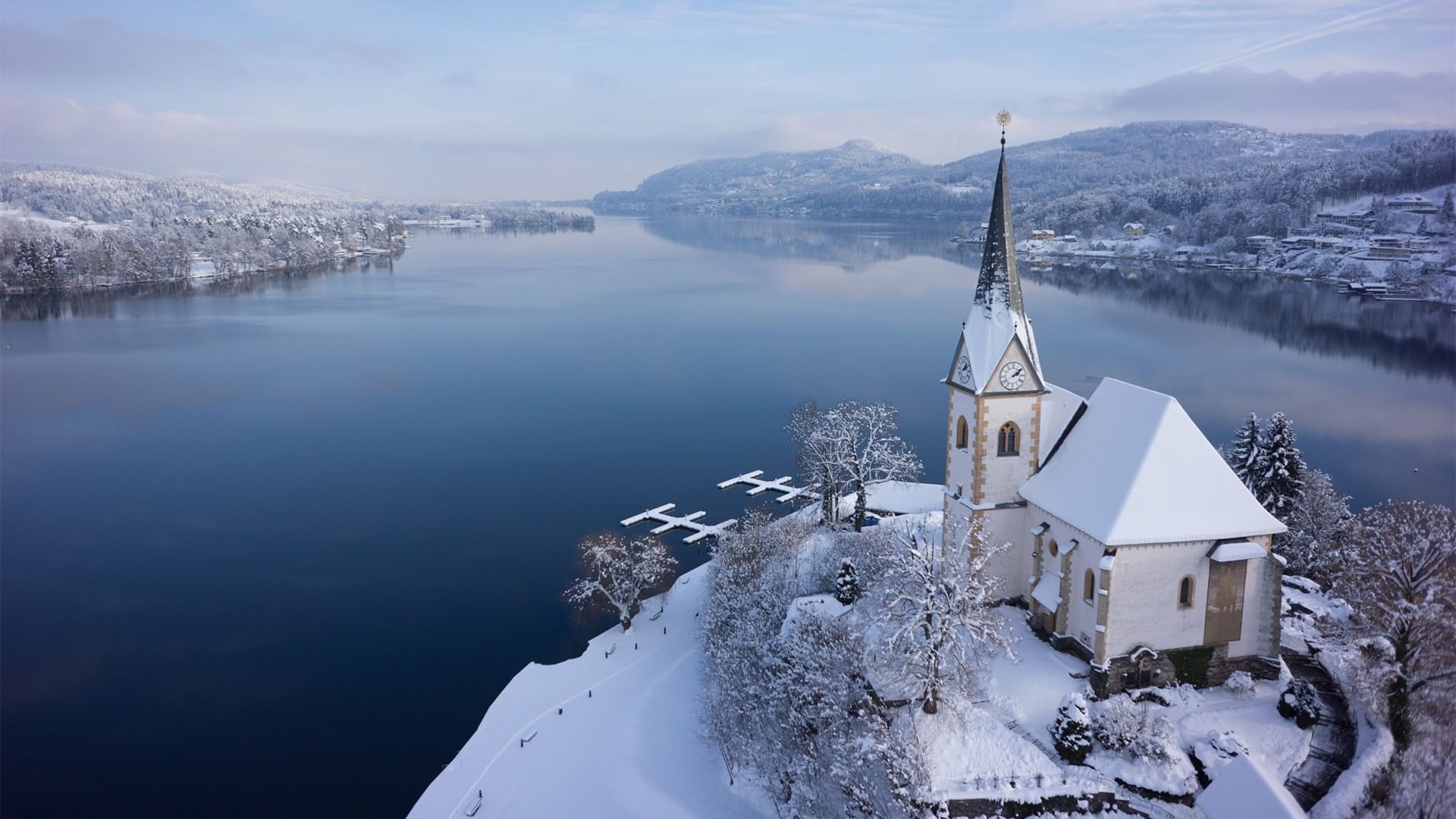 Snowy church by lake with mountains in background at Falkensteiner Schlosshotel Velden.