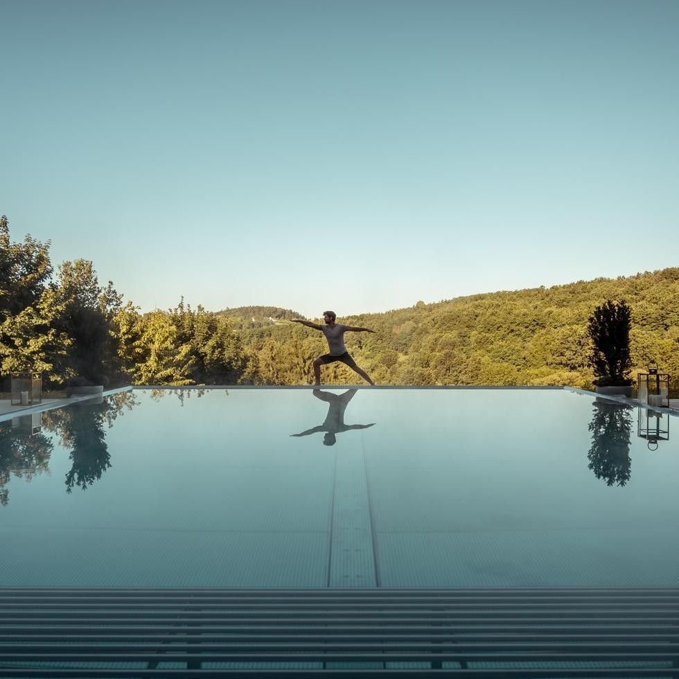 Person in Yogastellung über dem Pool, umgeben von Bäumen und Hügeln bei Yoga & Dance mit STUDIO herzfeld.