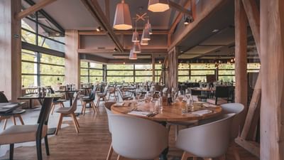 An elegant dining room with wooden floors, large windows, round tables, and modern lighting.