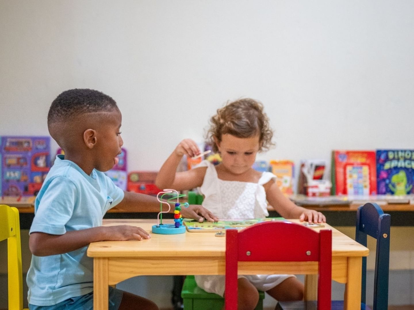 Two children playing with toys at a small table in a room with bookshelves.