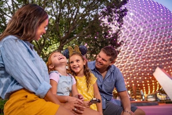 Two girls sit between a man and a woman in front of a tree and the Spaceship Earth orb at EPCOT. 