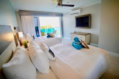 Two beds with blue towels and bags facing wall-mounted TV in Ocean Front Grand 2 Bedroom Suite at The Abidah Hotel by Accra