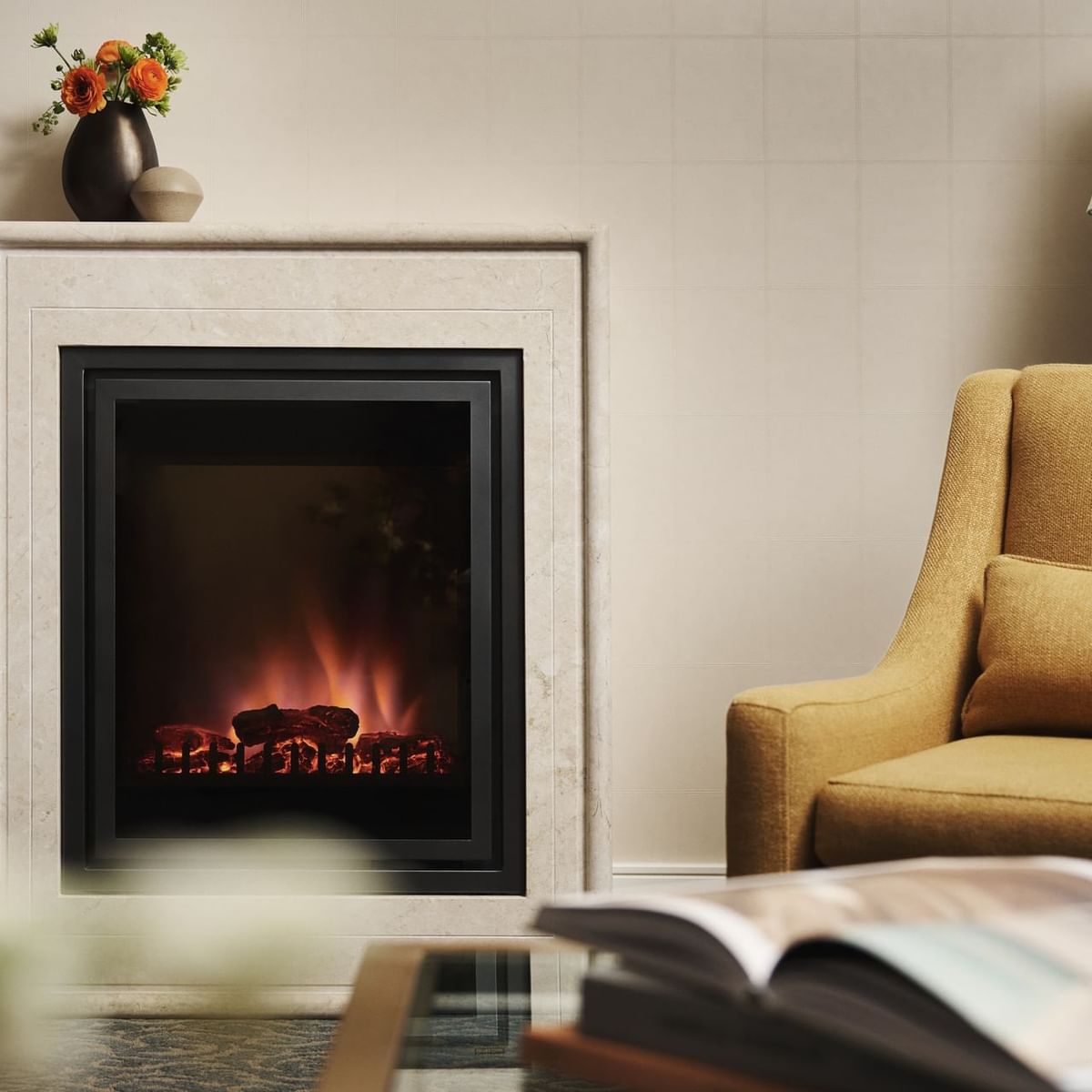 Fireplace with glowing embers, vase of flowers, armchair, and open book on a table in a room.
