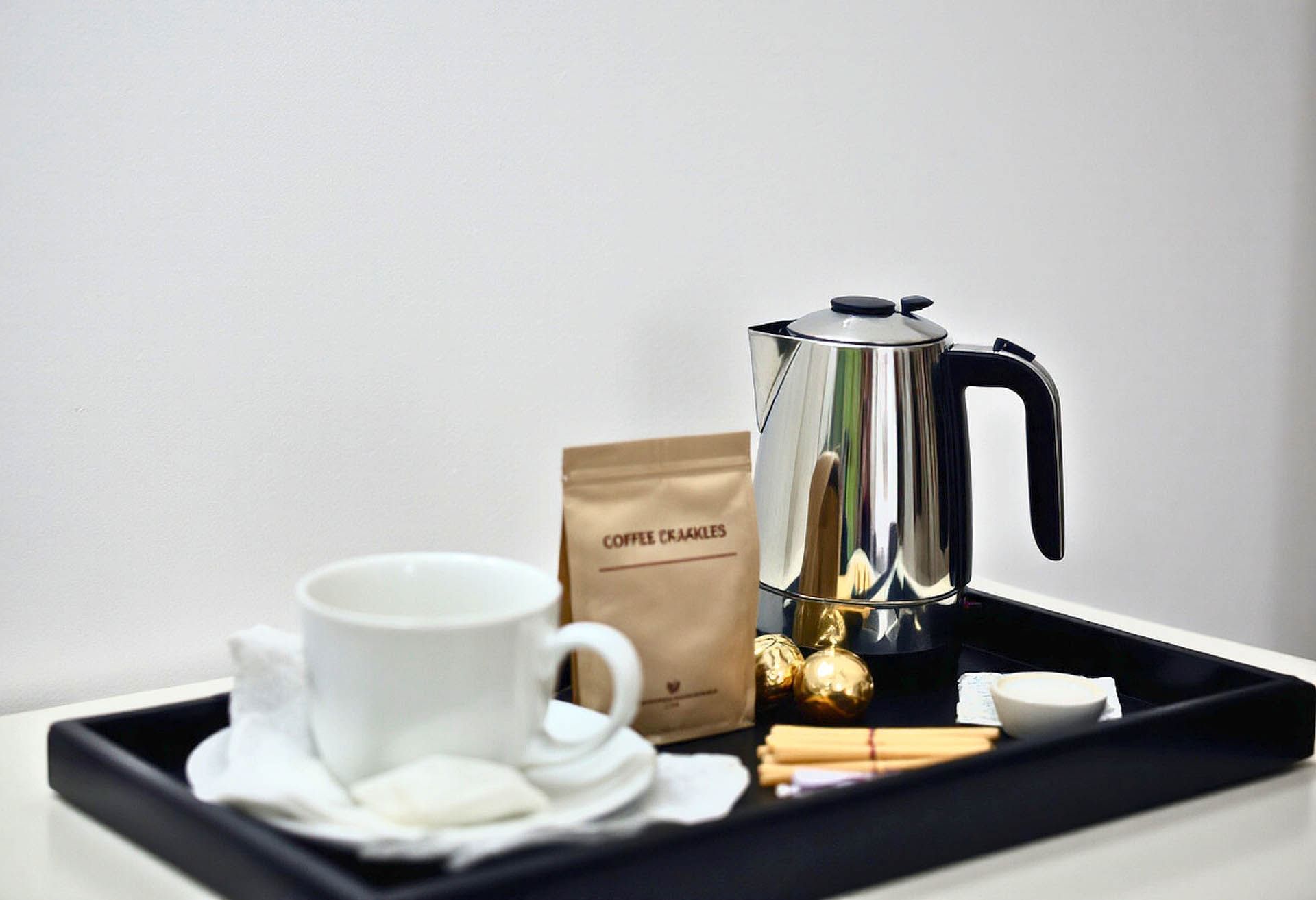 Silver coffee pot on a tray with a white cup, coffee packet, golden treats, and wooden stirrers at Avaneo Hotel