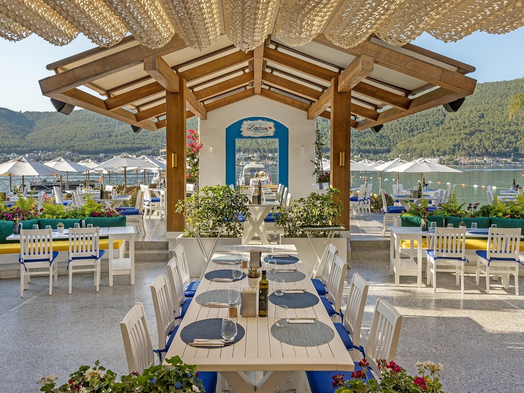 An open-air restaurant at Titanic Luxury Collection Bodrum with sea and mountain views, featuring a long white table and chairs.
