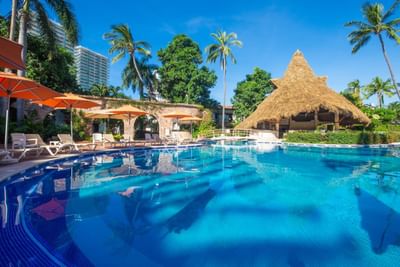 Resort pool with lounge chairs, umbrellas, palm trees, and a thatched-roof hut under a clear blue sky.