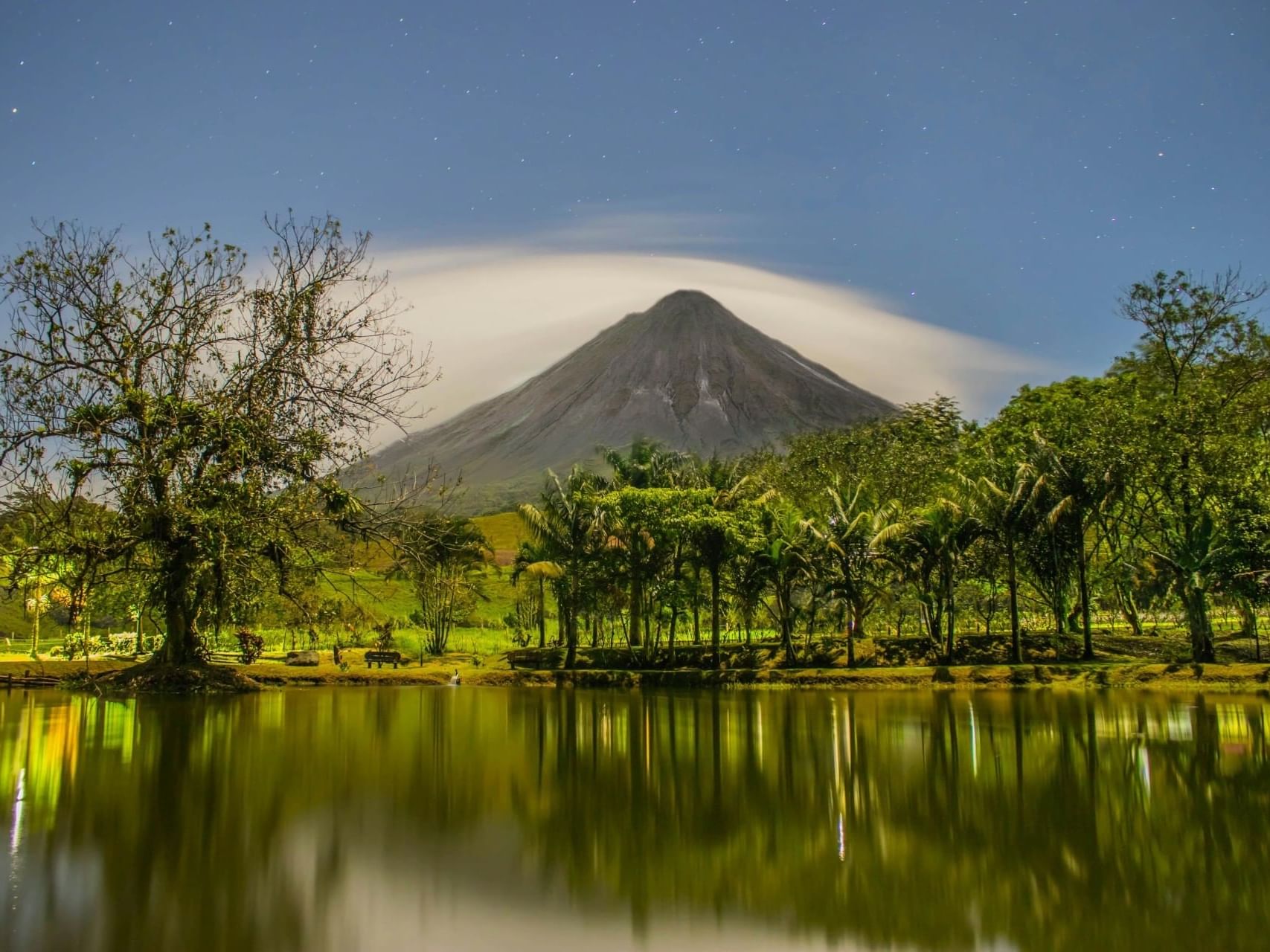 Distance view of Arenal Volcano reflected in a calm lake and trees in the foreground near El Silencio Lodge and Spa