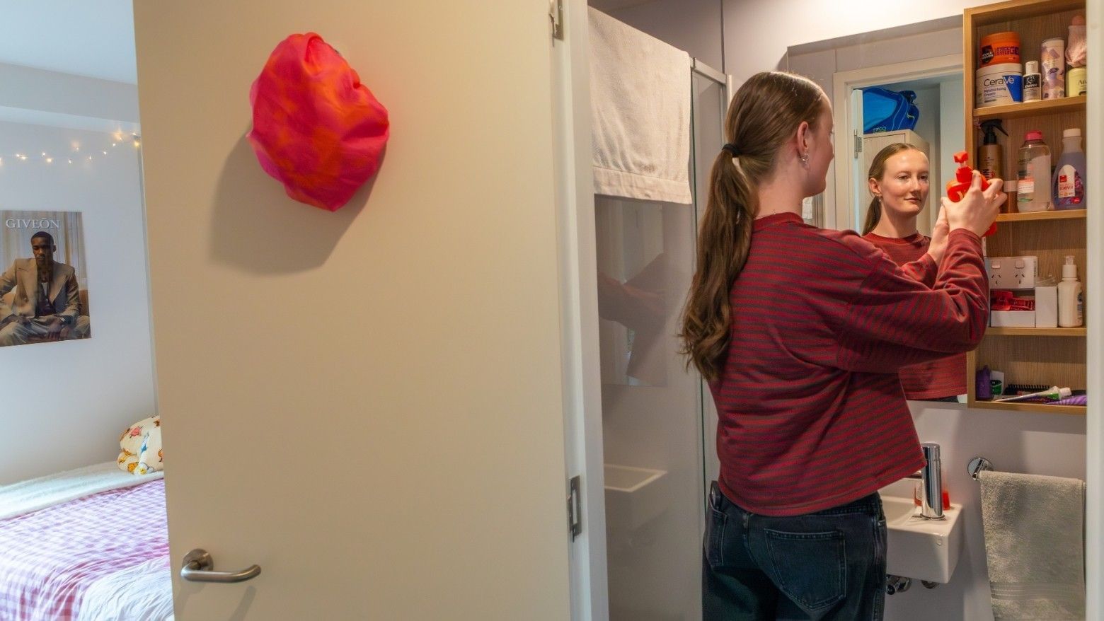 Woman in striped shirt reaching into bathroom cabinet with mirror, bedroom visible through open door with red wall art.