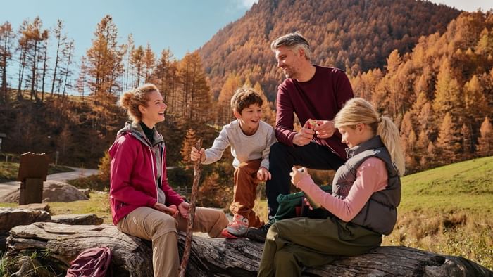 Family sitting on a wooden log under autumn trees near a green hill at Falkensteiner Hotels & Residences